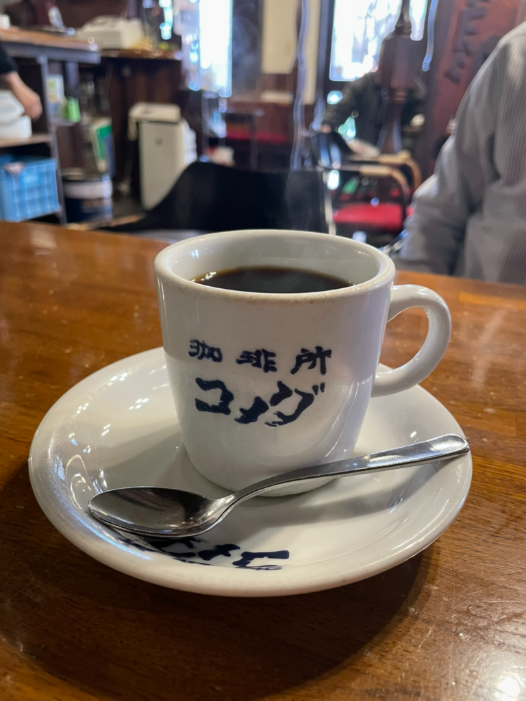 木目の美しいテーブルの上に置かれた、オールド・コメダのロゴが入った白いマグカップと熱いコーヒー、スプーンが乗ったソーサーのクローズアップ写真。