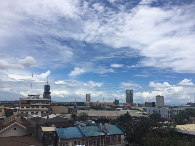 青い空と白い雲の下に広がる、フィリピンの街並み。手前には住宅の屋根、奥にはビルが見える。