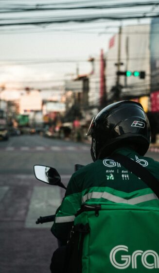 バイクに乗ったGrab Foodの配達員の背中。緑色の制服とロゴ入りの配達バッグが見える。街の風景が背景にある。