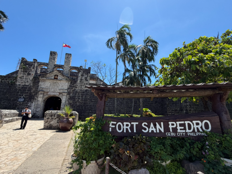 快晴の下に佇む石造りのサンペドロ要塞の入り口。手前には「FORT SAN PEDRO」と書かれた木製の大きな看板。