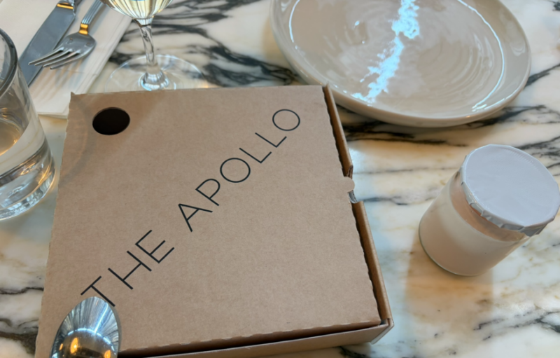 大理石のテーブルの上に置かれた、「THE APOLLO」のロゴが入ったクラフト紙のピタブレッド用テイクアウトボックス。隣には白いキャップ付きの小瓶があります。