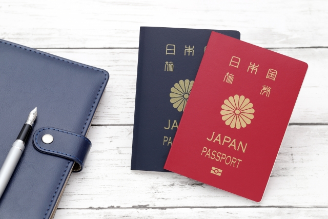 白い木目調のテーブルの上に置かれた、日本の紺色と赤色のパスポート、紺色の手帳、銀色の万年筆。