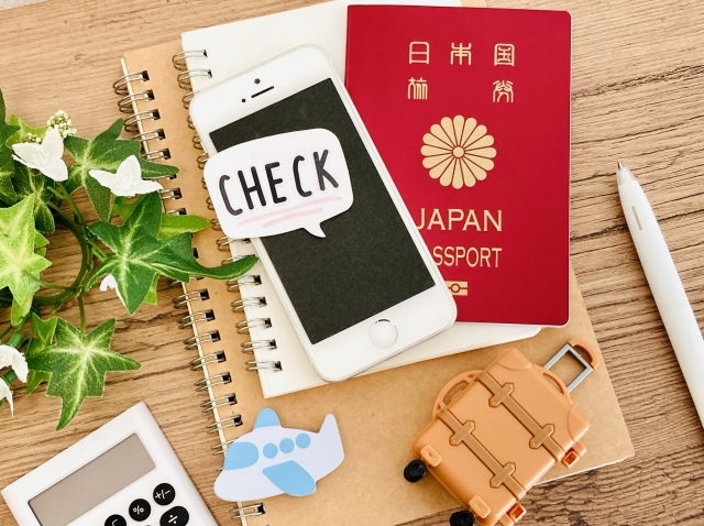 ノート、スマートフォン、パスポート、電卓、飛行機とスーツケースのイラストが描かれた、旅の準備・チェックリストをイメージした俯瞰写真。