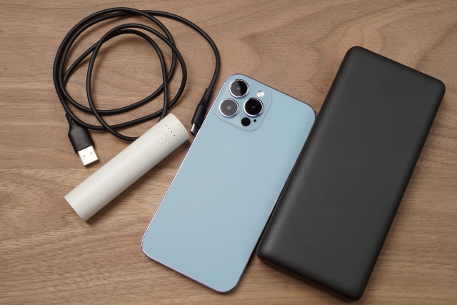 木目調のテーブルに置かれた水色のスマートフォン、黒い大容量モバイルバッテリー、白い小型バッテリー、充電用USBケーブル。