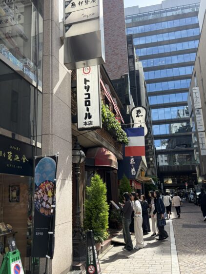 銀座の街角にあるトリコロール本店の看板。白い看板に黒い文字で店名が書かれ、隣にはフランス国旗が掲げられている。