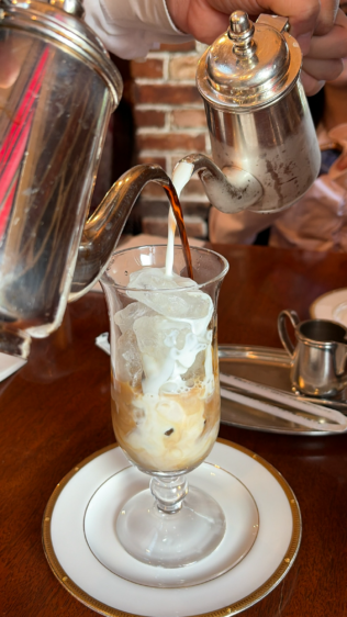 スタッフが2つの銀のポットから、氷の入ったグラスにコーヒーとミルクを同時に高い位置から注ぎ入れている瞬間。