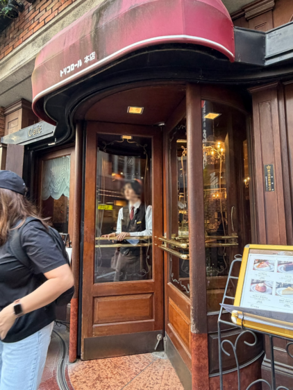 トリコロール本店の入り口。赤いオーニングの下に木製の回転扉があり、制服を着たスタッフが客を待っている様子。