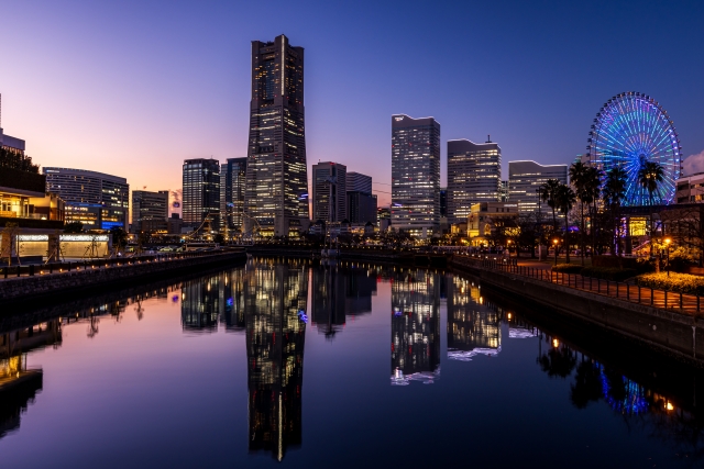 夕暮れから夜にかけてのグラデーションの空に浮かび上がる、横浜みなとみらいのビル群。横浜ランドマークタワーやカラフルにライトアップされた大観覧車(コスモクロック21)が、静かな水面に美しく反射している。