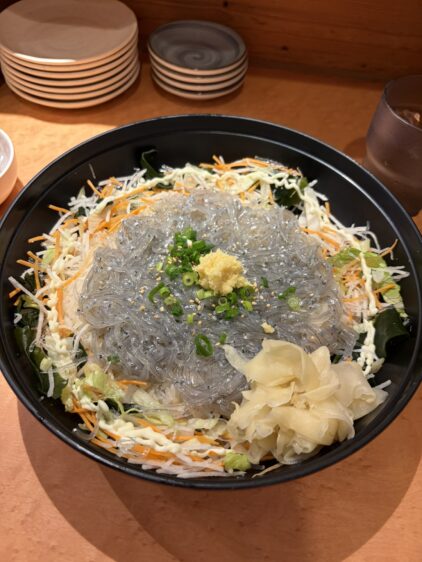 大きな黒い器に敷き詰められた千切り野菜の上に、たっぷりの生しらす、おろし生姜、刻みネギ、ごまがトッピングされた生しらす丼。