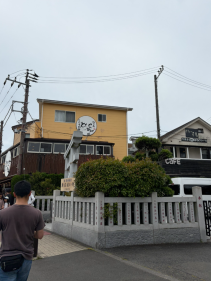 白い石鳥居の奥に見える、黄色い壁に「江の島 しらす問屋 とびっちょ」の丸いロゴ看板が掲げられた建物の外観。