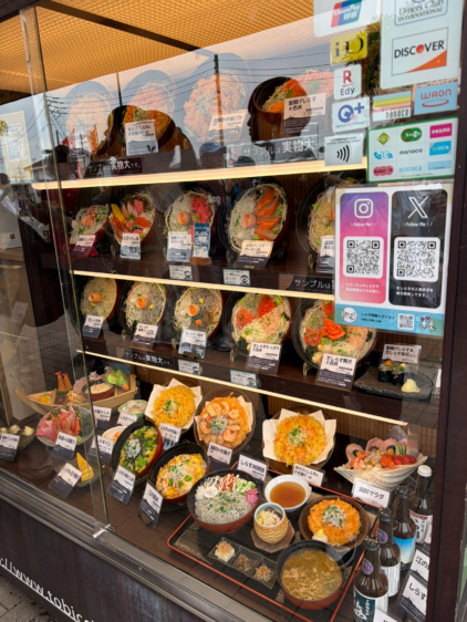 ガラスケースの中にずらりと並んだ、生しらす丼やかき揚げ丼などのリアルな実物大食品サンプルと、電子マネーの対応ステッカー。