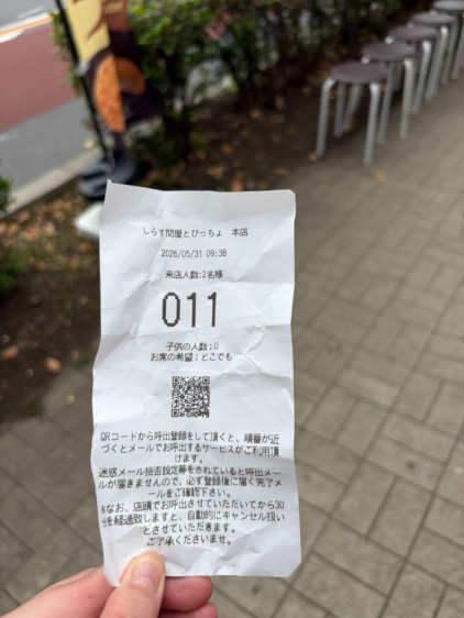 手に持った「しらす問屋 とびっちょ 本店」の整理券。番号は011、日時は2026年5月31日9時38分、来店人数2名と印字されている。