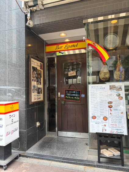 スペイン料理店「Bar Español」の入り口。木製のドアには「Open」の札が掛かり、左右にメニュー看板、ポスター、そして大きな生ハムが吊り下げられている。