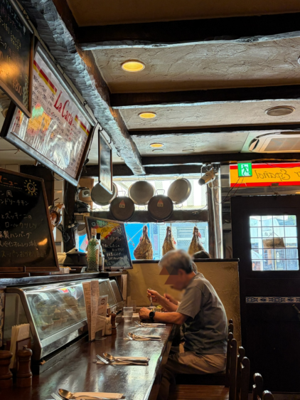 スペイン料理店の店内。木製のカウンター席に座り、食事をする男性。奥にはぶら下がった生ハム、調理器具、黒板メニューが見える。