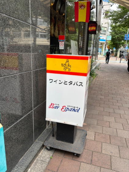 石畳の歩道に置かれた、スペイン国旗カラーの屋外看板。馬に乗った騎士のイラスト、「ワインとタパス」、「Bar Español」の文字が書かれている。背景には街並みと通行人が写っている。