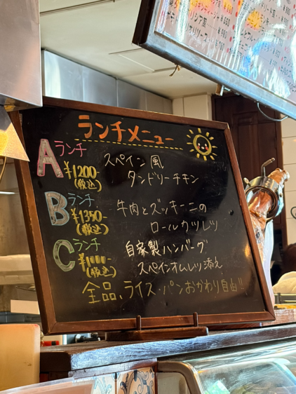 店内に置かれた手書きのランチメニュー黒板。A、B、Cのセット内容(スペイン風タンドリーチキン、牛肉とズッキーニのロールカツレツ、自家製ハンバーグ)と価格、ライス・パンおかわり自由であることが書かれている。