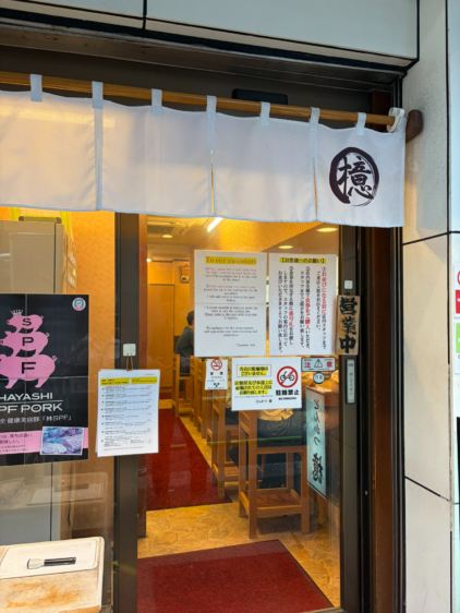 とんかつ檍の店構え。白い暖簾には店のロゴが描かれている。ガラス扉には日本語と英語の注意書き（To our customers、お客様へのお願い）や、「林SPFポーク」のピンク色のポスターが貼られている。