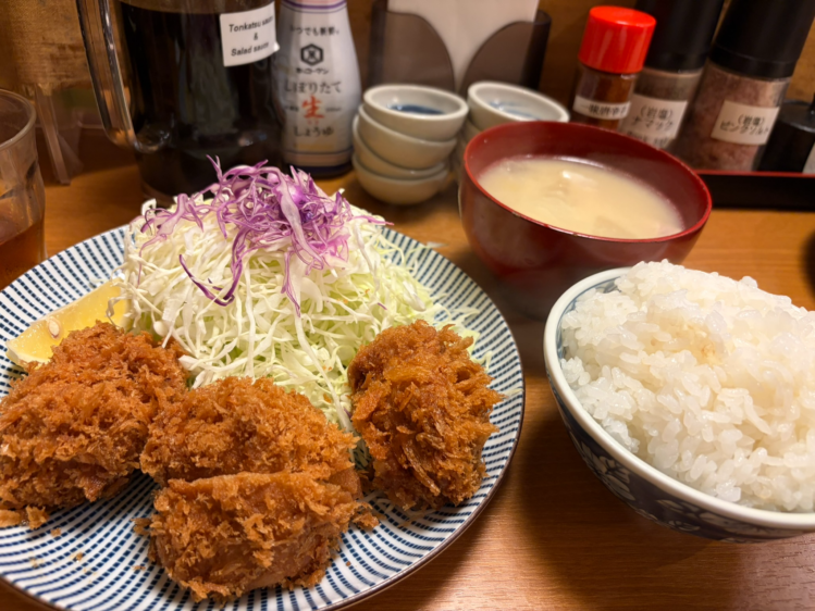木のカウンターに置かれたヒレカツ定食。手前から、ヒレカツとキャベツの皿、山盛りのご飯、具だくさんの豚汁、小鉢が並ぶ。背景には調味料がぼんやりと見える。