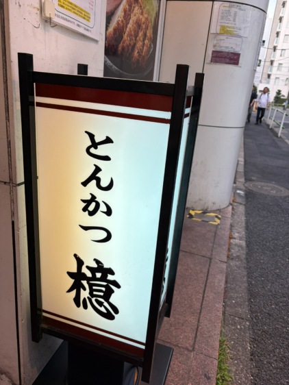 通りの角に立つ、ぼんやりと光る行燈看板。日本語で「とんかつ 檍」と書かれた大きな文字が目印。