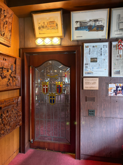 横浜スカンディヤの店内入口。中央にステンドグラスがはめ込まれた重厚な木製のドアがあり、両サイドの壁には大きな木彫りのレリーフが飾られている。ドアの上にはデンマークの風景画と店内のスケッチが額装されており、クラシカルな雰囲気が漂う。